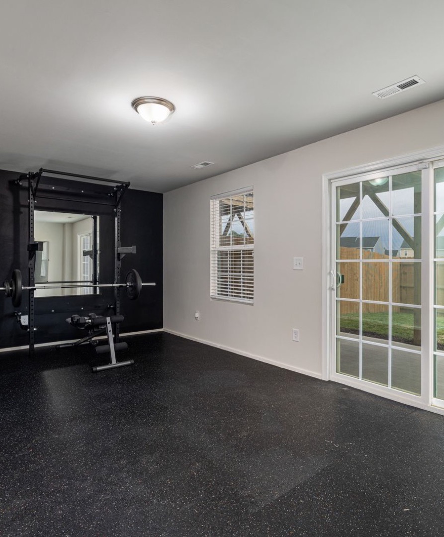 Bright gym room with large windows, wooden floor, black workout benches, and a mirror wall reflecting dumbbells and equipment.